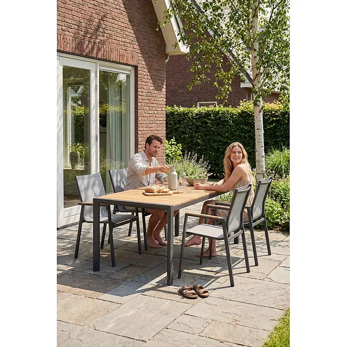 Barths Gartentisch Paros - L x B x H: 160 x 90 x 76 cm, Aluminium, Teak, Anthrazit/Braun - Outdoor-Essgruppe mit Holztisch und dunklen Metallstühlen mit hellgrauem Netzgewebe, von zwei Personen auf der Terrasse genutzt.