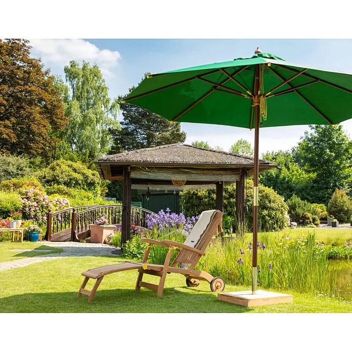 Kai Wiechmann Garten-Liegestuhl Teakholz Deckchair Brighton mit Rollen - Teak, Klappbar, Naturbelassen - Ein grüner Sonnenschirm und eine hölzerne Sonnenliege laden zum Entspannen in einem sonnigen Garten ein.