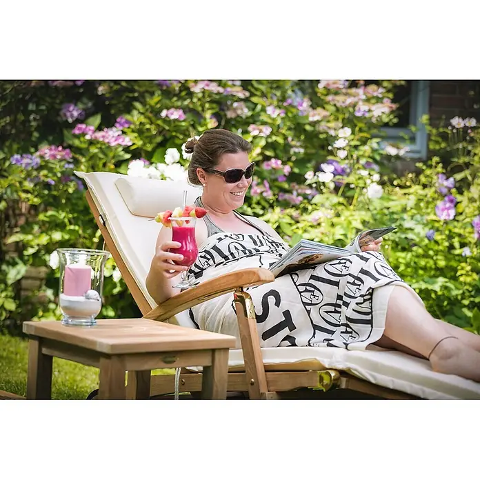 Kai Wiechmann Garten-Liegestuhl Teakholz Deckchair Brighton mit Rollen - Teak, Klappbar, Naturbelassen - Frau entspannt im Garten auf einer hölzernen Sonnenliege mit cremefarbenem Kissen, liest und genießt einen Drink.
