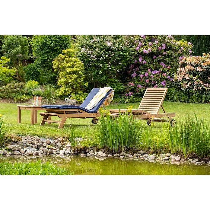 Kai Wiechmann Gartenliege Teak Sonnenliege Komfort - Naturbelassen, Holz, Rollbar - Zwei hölzerne Sonnenliegen, eine mit blauer Auflage, und ein kleiner Tisch in einem üppigen Garten an einem Teich.