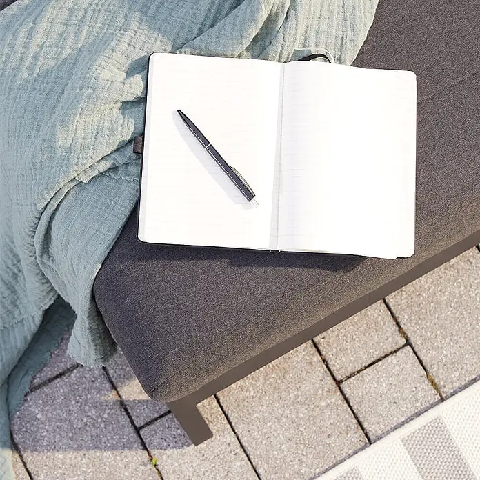 Home Deluxe Gartenliege VALENCIA - Grau, Sonstiges - Offenes Notizbuch mit Stift auf einem dunklen Loungesessel neben einer hellgrünen Decke.