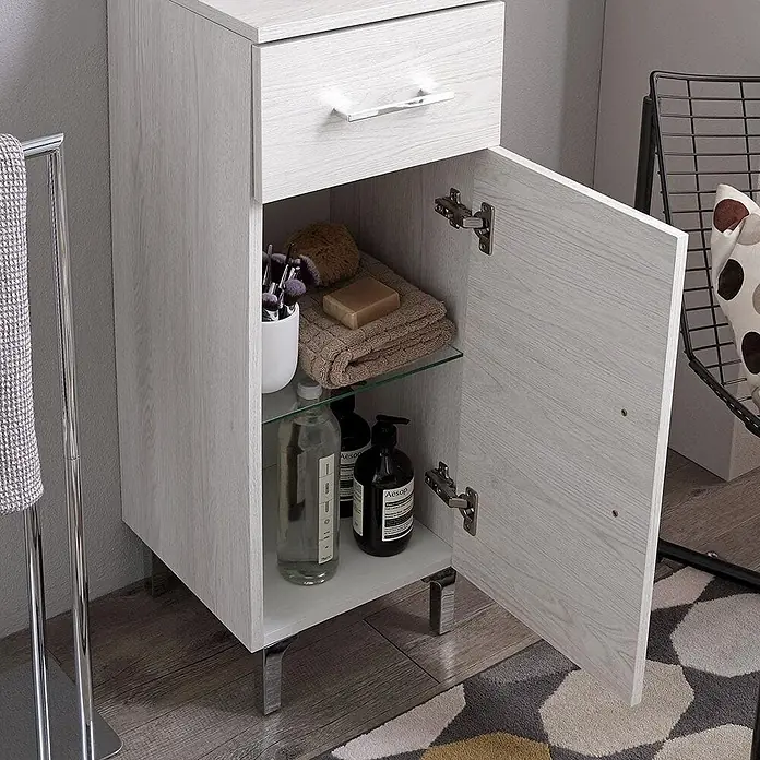 Kiamami Valentina Badmöbel-Set - Weiß - Hellgrauer Badezimmerschrank in Holzoptik mit Schublade und offenem Fach mit Glasablage, verchromten Griffen und Füßen, gefüllt mit Toilettenartikeln.