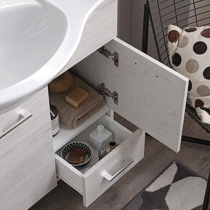 Kiamami Valentina Badmöbel-Set - Helles Holz - Eine Badezimmer-Waschtischanlage aus hellem Holz mit weißem Waschbecken, offener Tür und Schublade mit Toilettenartikeln.