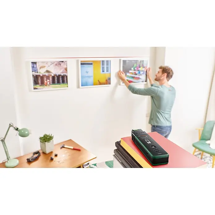 Bosch Laserentfernungsmesser Zamo Set - Messbereich: 0,15 - 40 m, 7 -tlg. - Grün-schwarzer Bosch Zamo Laserentfernungsmesser im Vordergrund auf Büchern, während Mann im Hintergrund Bilder aufhängt.