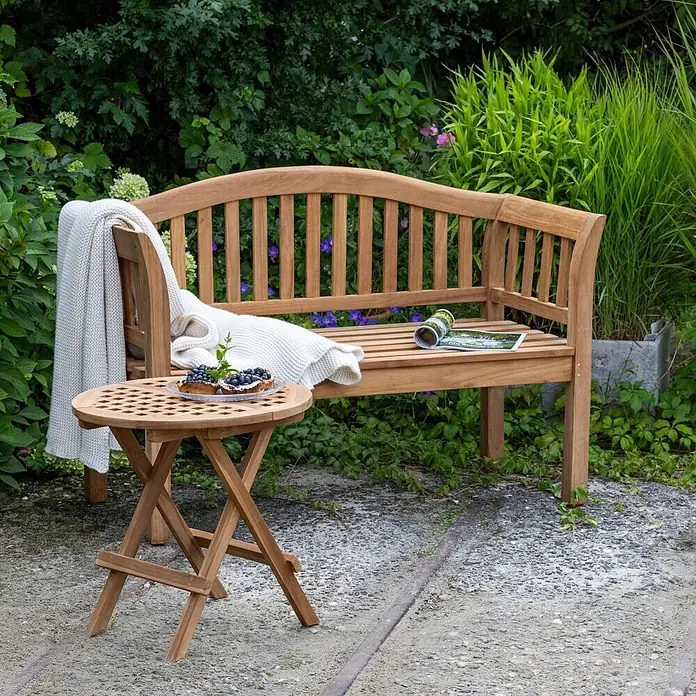 Bene Living Gartenbank Wave - Teakholz, Natur - Gartenbank und passender Klapptisch aus Naturholz in einer üppigen Gartenumgebung.