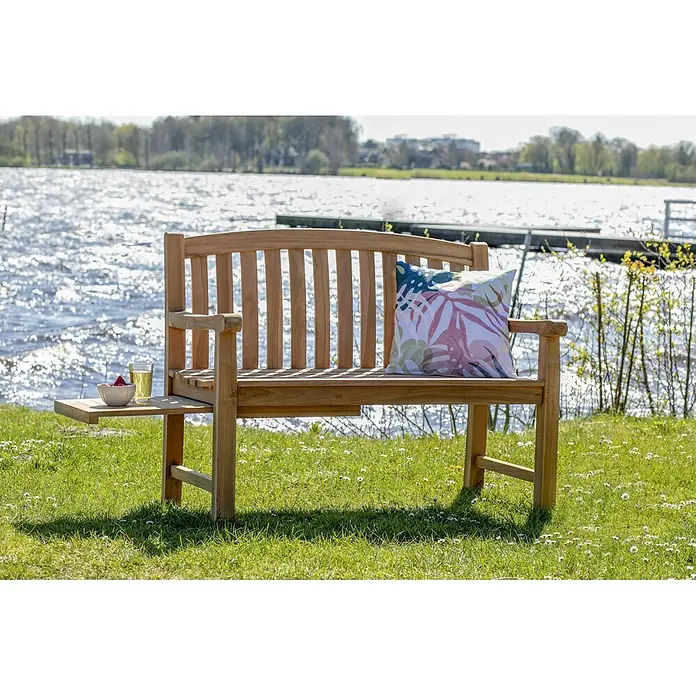 Bene Living Gartenbank Lancaster - Teakholz, Natur - Holzgartenbank mit ausziehbarer Seitenablage und gemustertem Kissen, auf Gras am Seeufer.