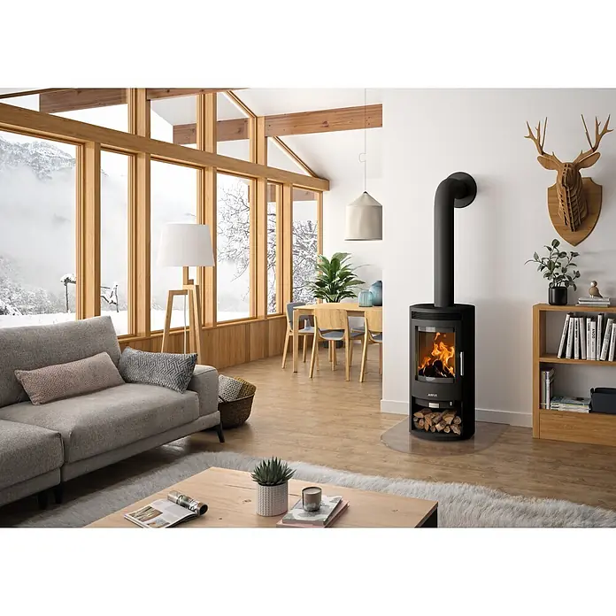 Justus Kaminofen Baltrum D 2.0 - 5,5 kW, Raumheizvermögen: 88 m³, Material Verkleidung: Stahl, Schwarz - Schwarzer Kaminofen mit sichtbaren Flammen und Holzscheitablage in einem modernen Wohnzimmer.