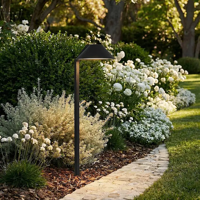 Lindby Erdspießleuchte Capelo -  - Eine moderne schwarze Wegbeleuchtung erhellt einen Steinweg im Garten, umgeben von Pflanzen und weißen Blumen.