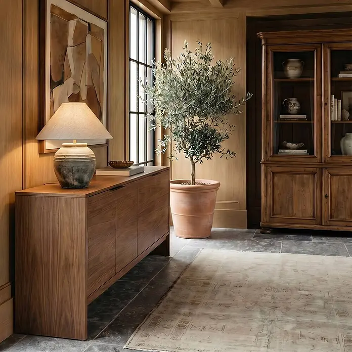 Lucande Tischleuchte Thalorin - braun / rost, E27 - Dunkles Holz-Sideboard mit Lampe und Büchern, platziert in einem Raum neben einem Fenster und Olivenbaum.