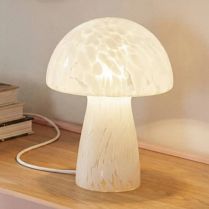 Lindby Tischleuchte Hubi - weiß / opal, weiß / opal, E27 - Pilzförmige weiße Tischlampe aus gesprenkeltem Glas, beleuchtet auf einem Holztisch neben Büchern.
