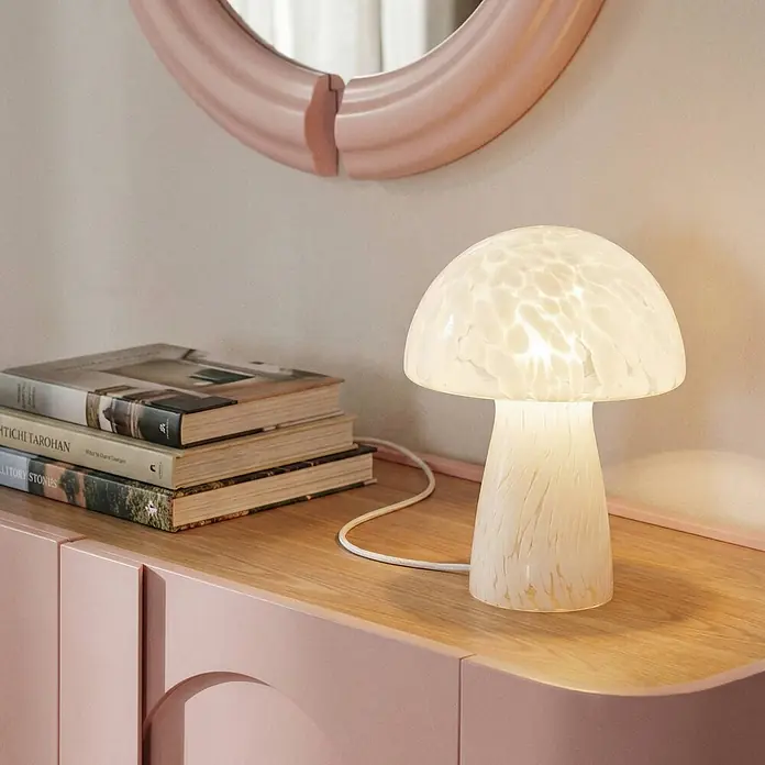Lindby Tischleuchte Hubi - weiß / opal, weiß / opal, E27 - Eine pilzförmige Tischlampe aus milchig-weißem gesprenkeltem Glas, die warmes Licht auf eine Holzoberfläche wirft.