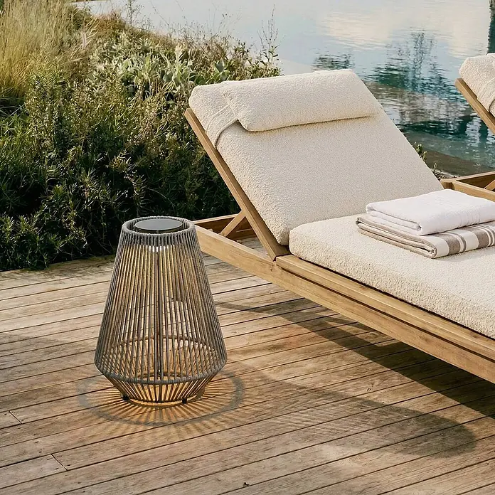 Lindby Außenwand- & Deckenleuchte Austas - alu / grau / zink, IP54 - Eine graue, konische, geflochtene Laterne leuchtet und wirft Lichtmuster auf eine Holzterrasse neben einer Sonnenliege, was eine einladende Outdoor-Atmosphäre schafft.