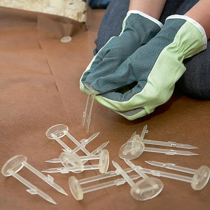 Dupont Plantex Befestigung Bio Pin - 12 Stk. - Person mit Handschuhen, die transparente Bodengewebeanker hält. Perfekt, um das Gewebe sicher am Boden zu befestigen.
