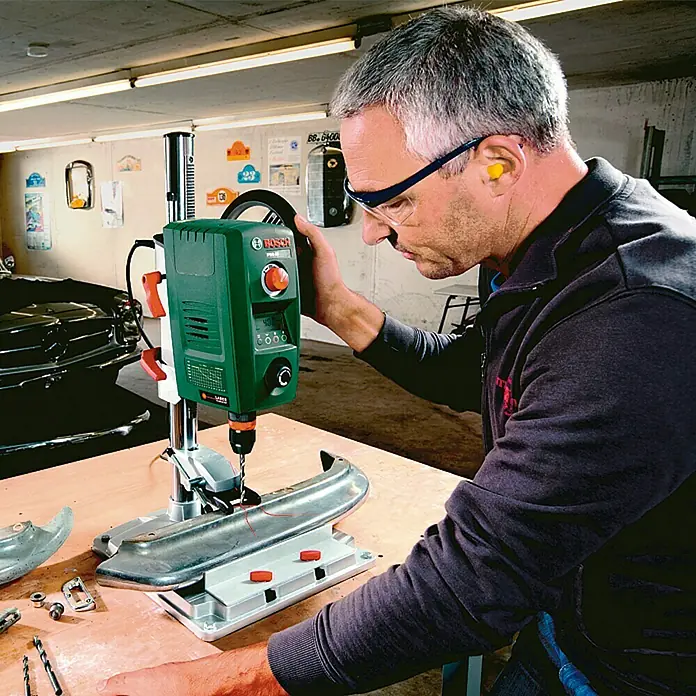 Bosch Tischbohrmaschine PBD 40 - Hub: 90 mm - Ein Mann bohrt mit einer Bosch PBD 40 Tischbohrmaschine ein Werkstück. Die Maschine ist grün und hat einen roten Knopf.