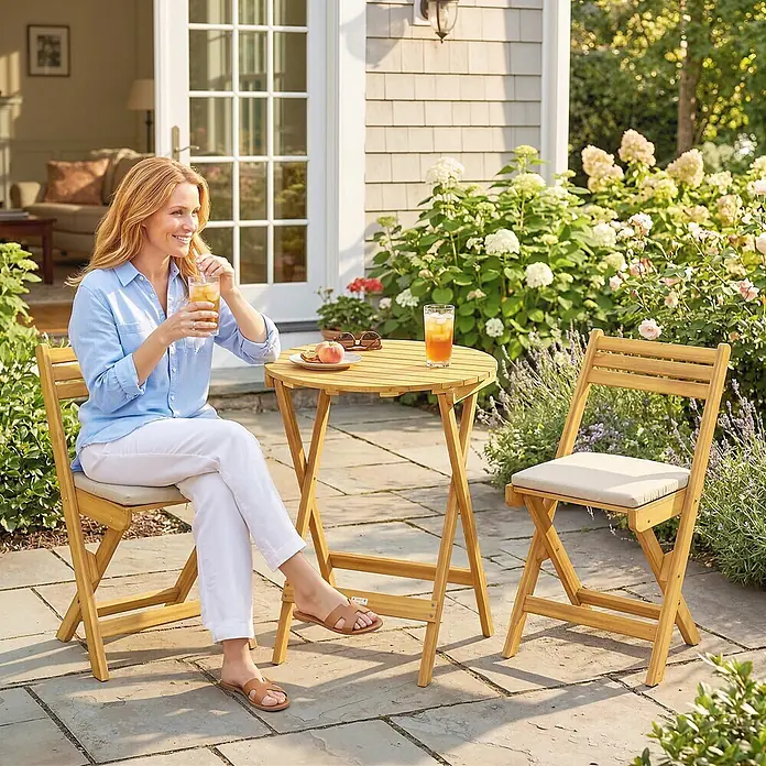 Outsunny Gartentisch-Set - Gesamtumfang: 3 -tlg., Akazienholz, Polyester, Schaumstoff, PP-Baumwolle, Klappbar - Helles Holz-Gartenmöbel-Set, bestehend aus Tisch und zwei Stühlen mit Kissen, genutzt von einer Frau, die ein Getränk genießt.
