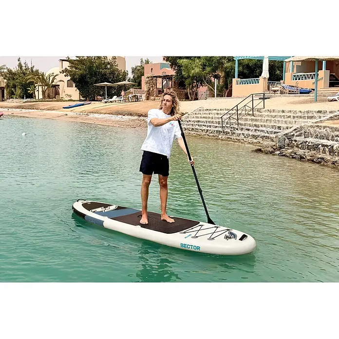 F2 SUP-Board-Set Sector "SMU 10,5" - L x B x H: 320 x 85 x 15 cm, Aufblasbar, Grau/Weiß - Mann beim Stand-up-Paddling auf einem weißen und blauen 'SECTOR' SUP-Board mit schwarzem Deckpad in ruhigem türkisfarbenem Wasser.