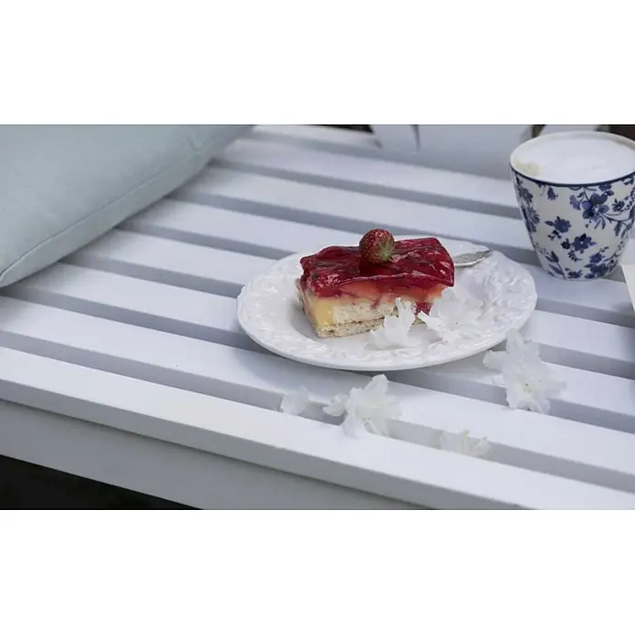 Bene Living Gartenbank Föhr - B x T x H: 130 x 53 x 83 cm, Mahagoni, Weiß - Ein Stück Erdbeerkuchen auf einem Teller, eine blaue Blumenkaffeetasse und ein helles Kissen auf einem weißen Lamellentisch.