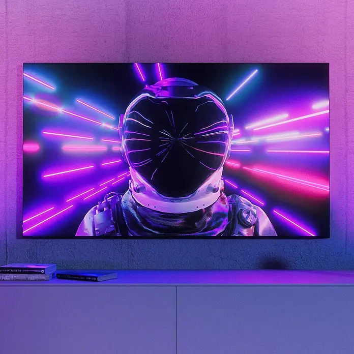 Philips Hue LED-Tischleuchte Play Wallwasher - L x B x H: 9,1 x 7,6 x 15,7 cm, Weiß, RGBW - Ein großer Bildschirm zeigt einen futuristischen Astronauten im Neonlicht, auf einem weißen Schrank in einem lila beleuchteten Raum.