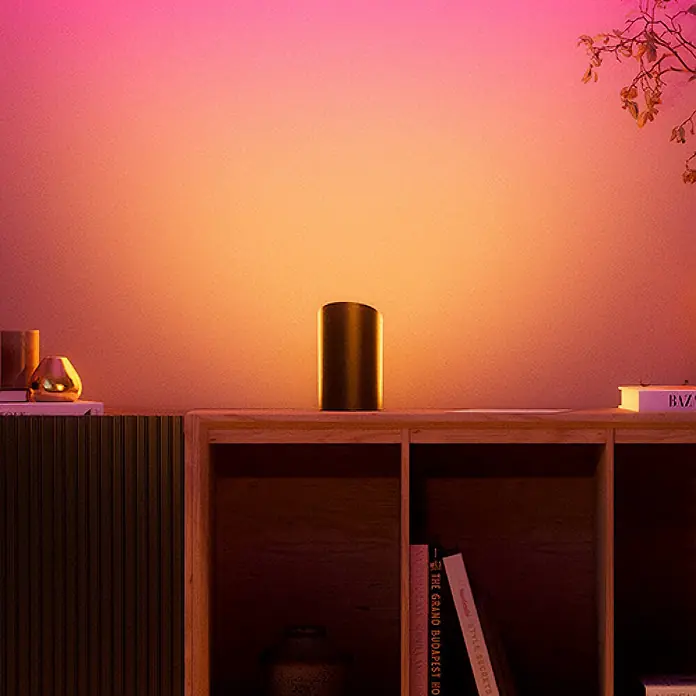 Philips Hue LED-Tischleuchte Play Wallwasher - L x B x H: 9,1 x 7,6 x 15,7 cm, Weiß, RGBW - Eine dunkle, zylindrische Lampe auf einem Holzregal beleuchtet die Wand mit einem leuchtenden Orange-Rosa-Glanz.