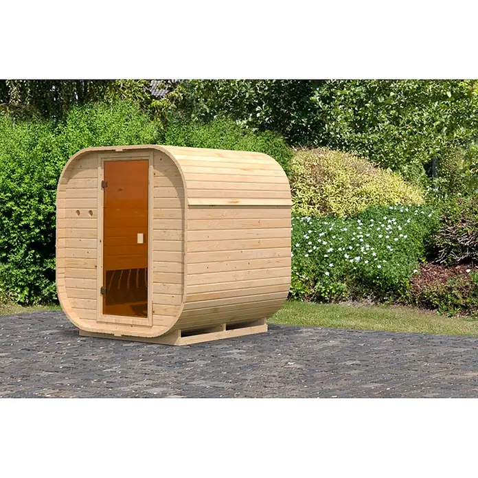 Karibu Saunahaus Quadro 1 - Ohne Ofen, Natur, L x B x H: 175 x 199 x 205 cm - Hölzerne Fasssauna mit bernsteinfarbener Glastür auf einer gepflasterten Terrasse im Garten.