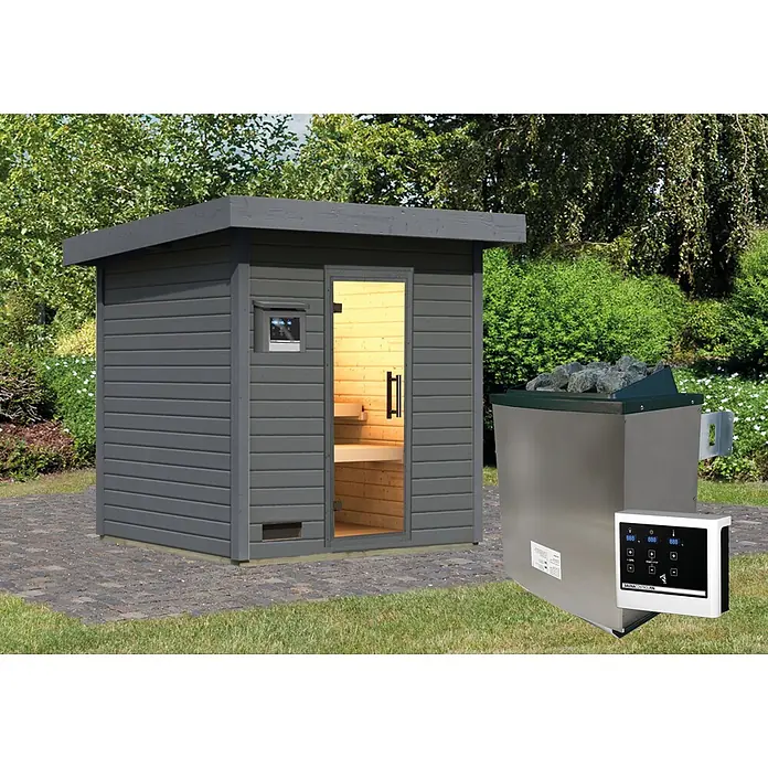 Karibu Saunahaus Kaapi 3 - 9 KW Ofen mit externer Steuerung, Anthrazit, L x B x H: 196 x 196 x 228 cm - Dunkelgraue Außensauna mit beleuchtetem Innenraum und Steuerfeldern, dazu ein Edelstahl-Saunaofen mit Steinen im Garten.