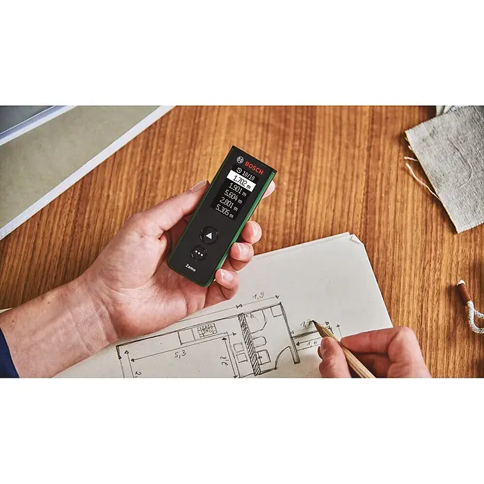 Bosch Laserentfernungsmesser Zamo Set - Messbereich: 0,15 - 40 m, 7 -tlg. - Hand hält grünes Bosch Zamo Lasermessgerät, zeigt Messwerte neben einem Grundriss.