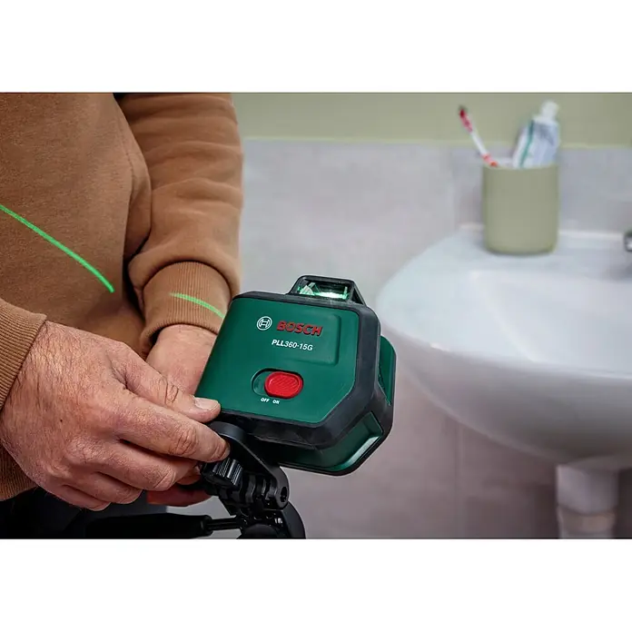 Bosch Kreuzlinienlaser PLL360-15G Set - Max. Arbeitsbereich: 30 m, 1 Akku, 3,6 V - Grünes Bosch PLL360-15G Laser-Nivelliergerät, von Händen gehalten, projiziert eine grüne Linie im Badezimmer.