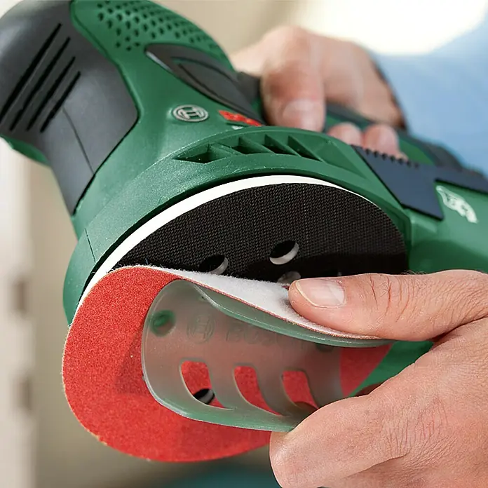 Bosch Elektro-Exzenterschleifer PEX 300 AE - 270 W, Durchmesser Schleifteller: 125 mm - Nahaufnahme eines grünen Bosch Prio Exzenterschleifers, der mit einer Hand bedient wird, um Schleifpapier zu wechseln.
