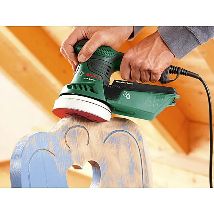 Bosch Lijadora excéntrica eléctrica PEX 300 AE - 270 W, Diámetro disco de lija: 125 mm - Una lijadora orbital Bosch PEX 300 AE en uso sobre una silla pintada en el interior de una casa con paneles de madera.