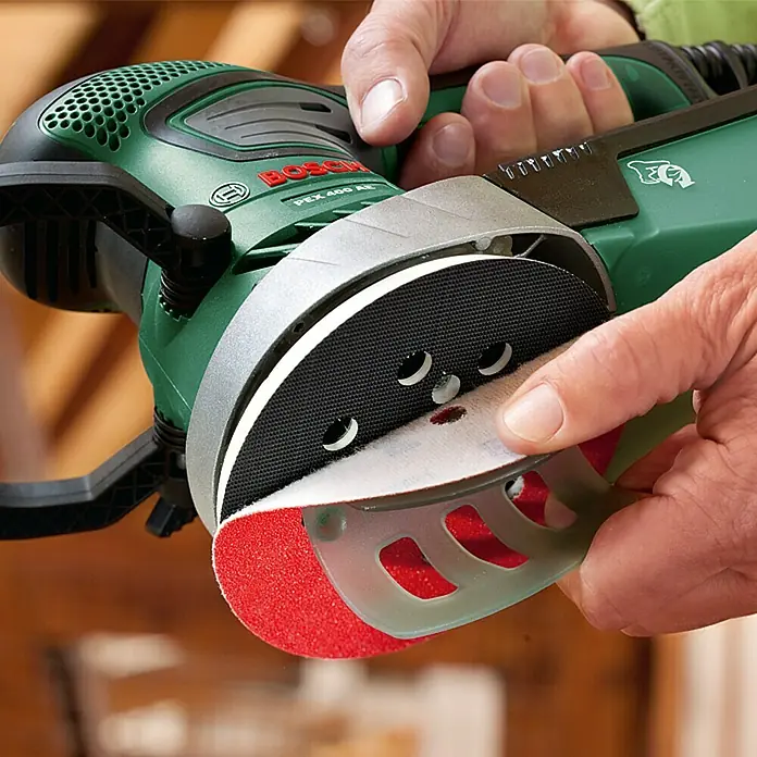 Bosch Exzenterschleifer PEX 400 AE - 350 W, Durchmesser Schleifteller: 125 mm - Nahaufnahme eines Bosch PEX 400 AE Exzenterschleifers beim Wechseln des Schleifpapiers, das rote Schleifmittel ist gut sichtbar.
