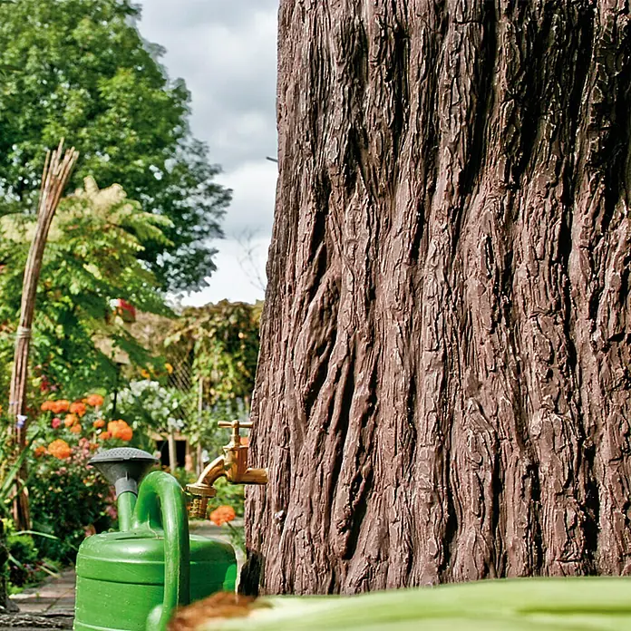Arves Regenspeicher Evergreen - 475 l, Dunkelbraun, 1 Stk. - Nahaufnahme eines dekorativen Wasserhahns an einem Baumstamm, daneben eine grüne Gießkanne, eingebettet in einen üppigen Garten.