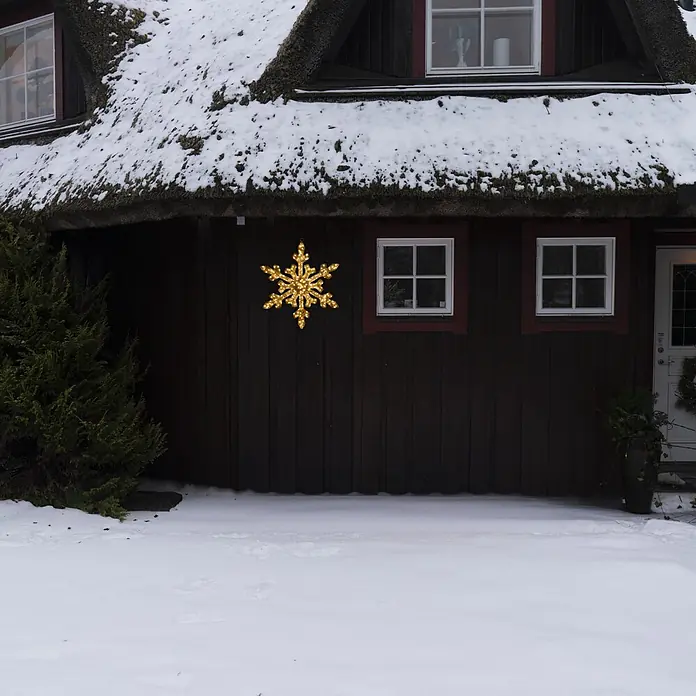 Konstsmide LED-Weihnachtsdekoleuchte Schneeflocke - 2,5 x 35 x 35 cm, Transparent, Netzbetrieben, LED - Eine leuchtende goldene Schneeflocken-Dekoration beleuchtet eine dunkle Holzwand eines schneebedeckten Hauses.