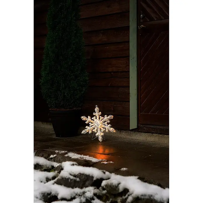 Konstsmide LED-Weihnachtsdekoleuchte Schneeflocke - 2,5 x 35 x 35 cm, Transparent, Netzbetrieben, LED - Leuchtende weiße Schneeflocken-Weihnachtsdekoration mit warmen Lichtern, stehend auf nassem Boden neben Pflanze und Holzgebäude.