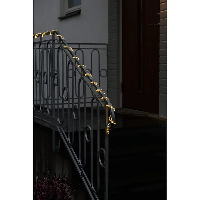 Konstsmide LED-Lichtschlauch Mini - Länge: 5 cm, Lichtfarbe: Warmweiß, 3,6 W, 392 lm, 220 V - 240 V - Warmweiße Lichterkette um ein dunkles Metallgeländer an einer Außentreppe, die zu einer Haustür führt.