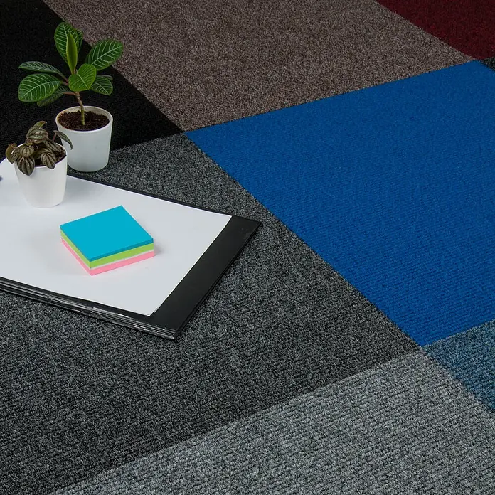 Nadelvlies Teppich Fliesen grau Selbstliegende Teppichfliesen in 50x50 cm aus Nadelfilz Schatex Teppichbodenfliesen dunkelgrau ideal für Büro -  - Bunte modulare Teppichfliesen in Grau, Blau, Braun und Rot, arrangiert mit Büroartikeln wie Pflanzen und Haftnotizen.
