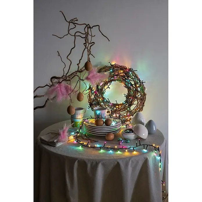 Konstsmide LED-Clusterlichterkette Pastell - Außen, Länge: 8,78 cm, 400 Stk., Netzbetrieben - Ostertischdekoration mit Ästen, hängenden Eiern, Federn, einem beleuchteten Kranz, Tellern und Schokoladeneiern.