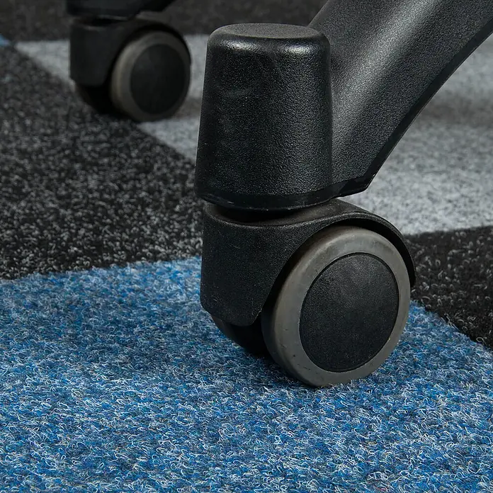 Bricoflor Teppichfliese - Breite: 50 cm, Nadelfilz, Polypropylen, blau - Nahaufnahme einer schwarzen Lenkrolle aus Kunststoff mit grauer Gummilauffläche auf einem blau, grau und schwarz gemusterten Teppich.
