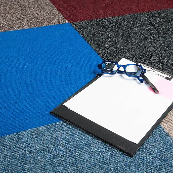 Bricoflor Teppichfliese -  - Blaue, achteckige Anne & Valentin Brille liegt auf einem Klemmbrett mit Papier und Stift, auf einem bunten Teppich.
