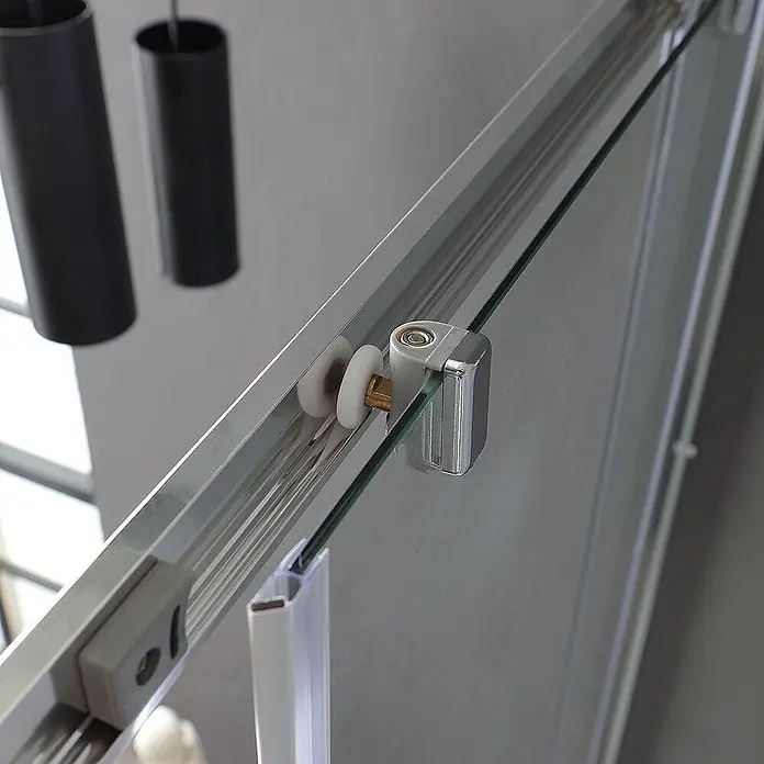 Kiamami Valentina Eckdusche - 700 x 1.000 x 1.850 cm, Transparent, Stärke: 4 mm - Silberner Schiebetürrollenmechanismus mit weißen Rädern an einer klaren Glasscheibe in einer Aluminiumschiene.