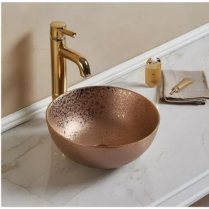 Sanicomfort Aufsatzwaschbecken Osiris - 35,8 x 35,8 cm, Hahnloch: Ohne, Ohne Überlauf, Rosegold - Ein rundes, metallic-kupferfarbenes Aufsatzwaschbecken mit texturierter Oberfläche und goldener Armatur auf einem weißen Marmorwaschtisch.