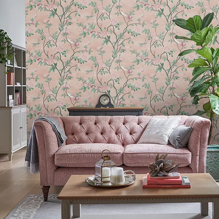 Laura Ashley Vliestapete Hagebuttenspur - Rosa, Floral, 1.000 x 52 cm - Rosafarbene Tapete mit zartem Blumen- und Vogelmuster, die eine Wohnzimmerwand hinter einem altrosafarbenen, getufteten Sofa schmückt.