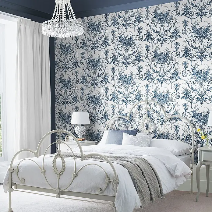 Laura Ashley Vliestapete Tuileries Französisch - Blau, Floral, 1.000 x 52 cm - Ein kunstvolles, cremefarbenes Metallbett mit weißer Bettwäsche, in einem Schlafzimmer mit auffälliger blau-floraler Tapete und Kronleuchter.