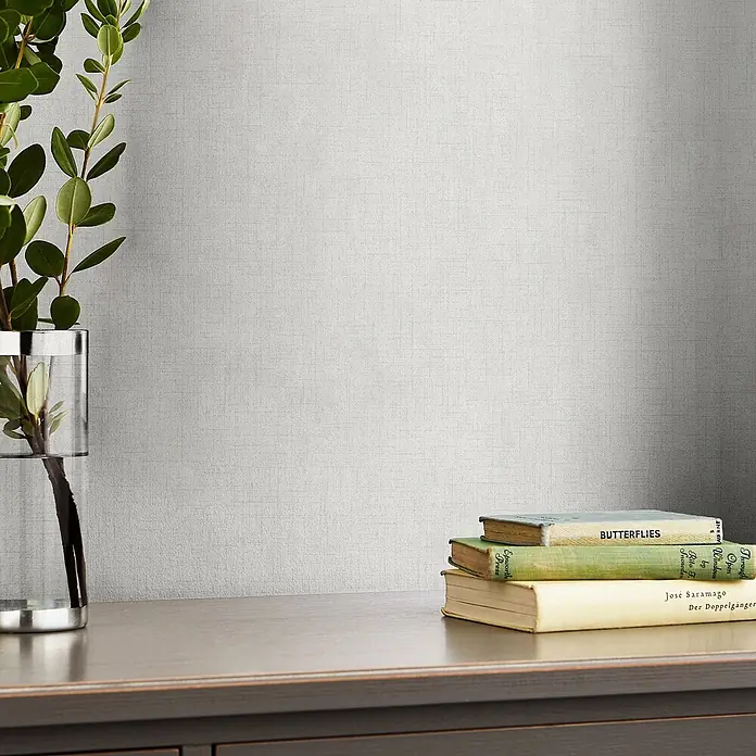 Laura Ashley Vliestapete Einfarbig - Silber, Uni, 1.000 x 52 cm - Hellgraue Tapete mit dezentem Webmuster, zu sehen in einer Raumszene hinter einer Pflanze und Büchern.
