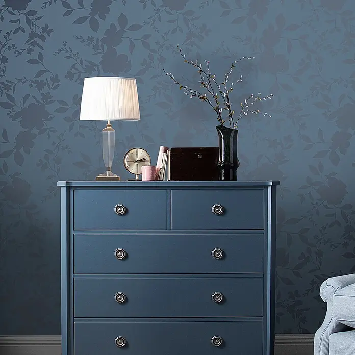 Laura Ashley Vliestapete Westbourne - Blau, Floral, 1.000 x 52 cm - Blaue Kommode mit silbernen Knöpfen, Lampe, Uhr, Büchern und Vase darauf, vor einer floral gemusterten blauen Wand.
