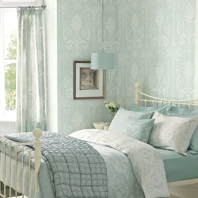 Laura Ashley Vliestapete Vliestapete Josette Damast - Blau, Ornament, 10 x 0,52 m - Hellblau-weißes Bettwäsche-Set mit Blumenmuster und gesteppter Decke auf einem Bett im Schlafzimmer.
