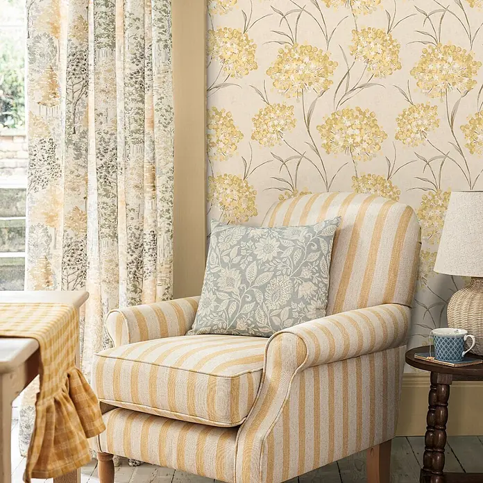 Laura Ashley Vliestapete Giselle Blumen - Gelb/Ocker, Floral, 1.000 x 52 cm - Gestreifter gelb-elfenbeinfarbener Sessel mit blauem Blumenkissen in einem gemütlichen Raum.