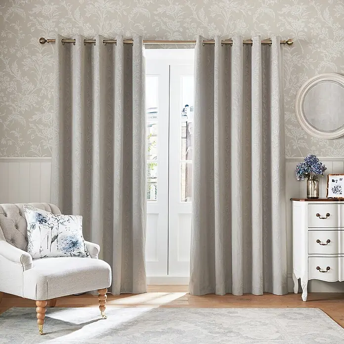 Laura Ashley Vliestapete Lloyd - Beige, Floral, 1.000 x 52 cm - Hellbeige Ösenschals mit dezentem Blumenmuster hängen an einer goldenen Stange und umrahmen eine Glastür.