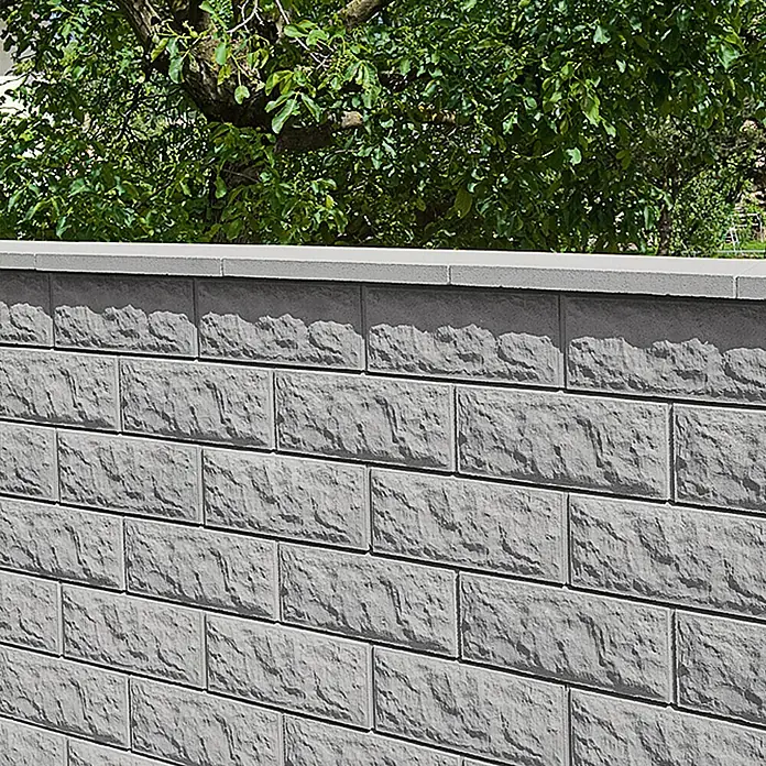 Mauerabdeckplatte Satteldach - Grau/Anthrazit, 50 x 30 cm, Beton - Graue Mauer aus Steinblöcken mit einer glatten Abdeckung, im Hintergrund grüne Bäume.