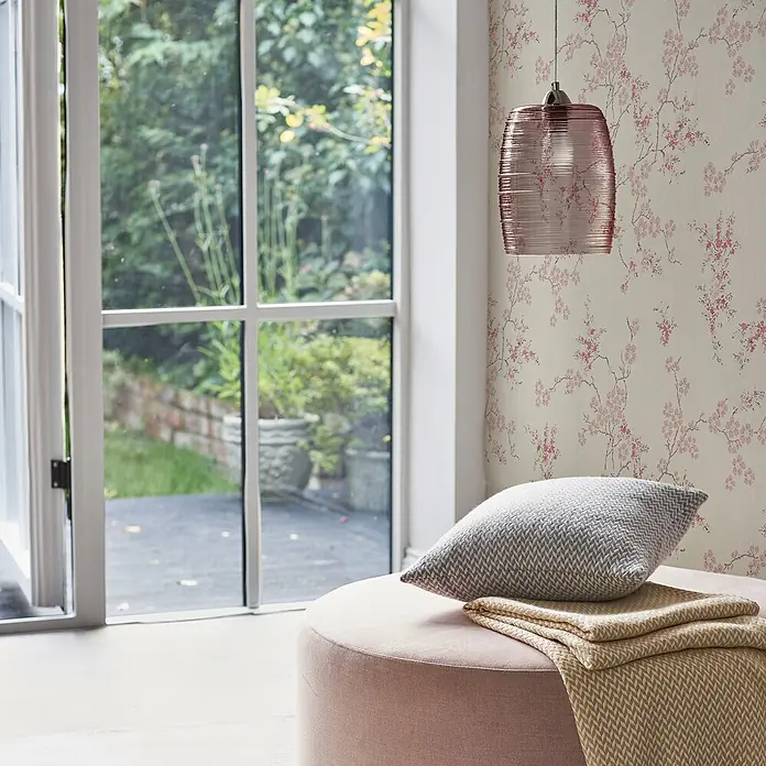 Laura Ashley Vliestapete orientalische Blüte - Rosa, Floral, 1.000 x 52 cm - Rosa gepolsterter Hocker mit grauem gemustertem Kissen und gelbem Überwurf, in einem Raum mit Blumentapete und rosa Pendelleuchte.
