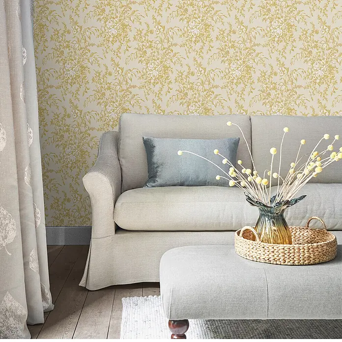 Laura Ashley Vliestapete Picardie - Gelb, Floral, 1.000 x 52 cm - Ein gemütliches Wohnzimmer mit hellbeigem Sofa, passendem Hocker mit geflochtenem Tablett und gelber Blumentapete.